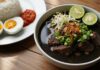 Resep Rawon Sapi Sederhana dengan Bumbu Rempah Lengkap Semangkuk Rawon Sapi hangat dengan taburan bawang goreng dan irisan jeruk nipis, disajikan dengan nasi dan telur rebus.